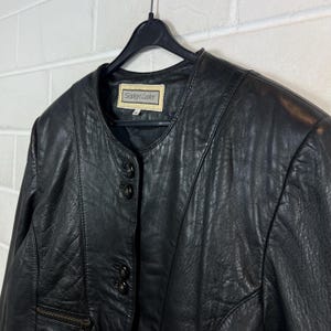 Op de afbeelding: Zwarte leren jas met een ronde kraag en drie knopen. De jas heeft een ritszak aan de linkerkant en het label luidt "Seiden Leder". De jas hangt aan een zwarte hanger.