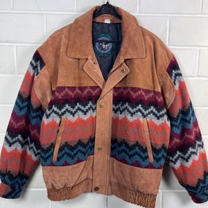 Könnte beinhalten: Braune Wildlederjacke mit einem Muster auf den Ärmeln und der oberen Brust. Die Jacke hat eine Knopfleiste, zwei Taschen und einen gerippten Saum. Das Muster umfasst rote, blaue und orangefarbene Zickzacklinien.