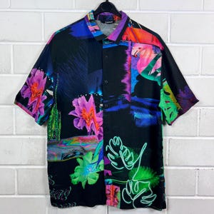 Pode incluir: Uma camisa de manga curta com um design floral e abstrato vibrante e colorido. A camisa apresenta um fundo preto com detalhes em verde néon, rosa, roxo e azul. A camisa está pendurada em um cabide preto.