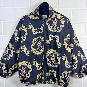 Pode incluir: Casaco preto com zíper com um padrão barroco dourado e detalhes rosa. O casaco tem uma gola alta e a marca "Slade" é visível. O casaco está pendurado contra uma parede de tijolos brancos.