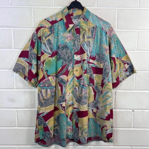 Könnte beinhalten: Kurzärmeliges Hemd mit farbenfrohem, abstraktem Muster. Das Hemd hat eine Mischung aus teal, bordeaux, gelben und grauen Formen. Das Hemd hängt an einem schwarzen Kleiderbügel.