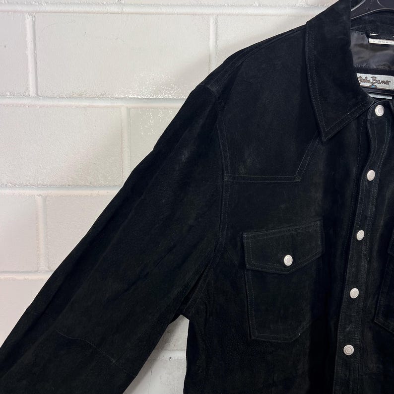 Peut inclure: Chemise en daim noir avec une fermeture boutonn&eacute;e, deux poches de poitrine et un col classique. La chemise a un d&eacute;tail de plastron de style occidental et des boutons blancs. Le tissu semble doux et a une finition mate.
