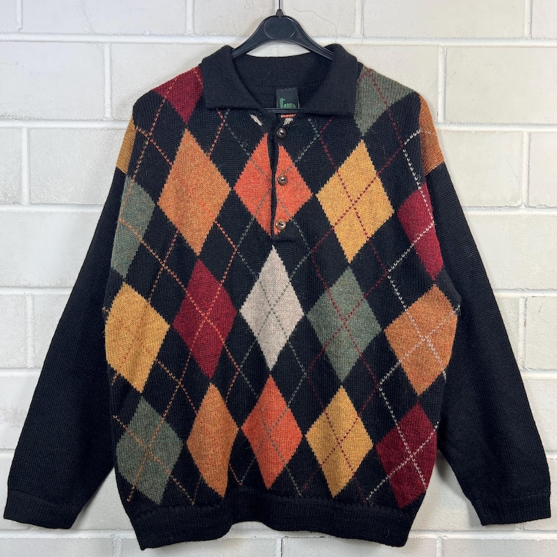 K&ouml;nnte beinhalten: Ein schwarzer Argyle-Pullover mit Kragen und drei Kn&ouml;pfen. Das Argyle-Muster zeigt Rauten in Orange-, Gelb-, Gr&uuml;n- und Rott&ouml;nen. Die &Auml;rmel und der Kragen sind einfarbig schwarz.