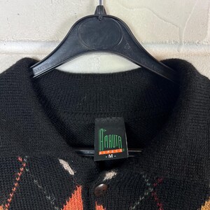 K&ouml;nnte beinhalten: Schwarzer Pullover mit Argyle-Muster und orangefarbenen und wei&szlig;en Akzenten. Der Pullover hat eine Knopfleiste vorne und einen Kragen. Das Etikett lautet "AMAUTA" in Gr&uuml;n und Wei&szlig;. Der Pullover h&auml;ngt an einem schwarzen Kleiderb&uuml;gel.