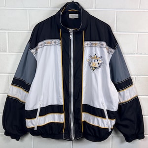 Könnte beinhalten: Eine schwarz-weiß-graue Reißverschlussjacke mit Stehkragen, langen Ärmeln und zwei Vordertaschen. Auf der Brust befindet sich ein dreieckiges Emblem mit dem Text OKC und Sternmotiven, während ein gemustertes Band über den Brustbereich verläuft.