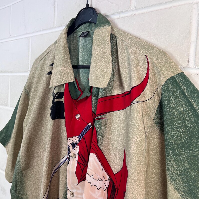 K&ouml;nnte beinhalten: Ein gemustertes Button-Down-Hemd in Beige, Gr&uuml;n und Rot. Das Design des Hemdes zeigt eine Figur mit einem Schwert. Das Hemd h&auml;ngt an einem schwarzen Kleiderb&uuml;gel. Es hat kurze &Auml;rmel und einen Kragen.