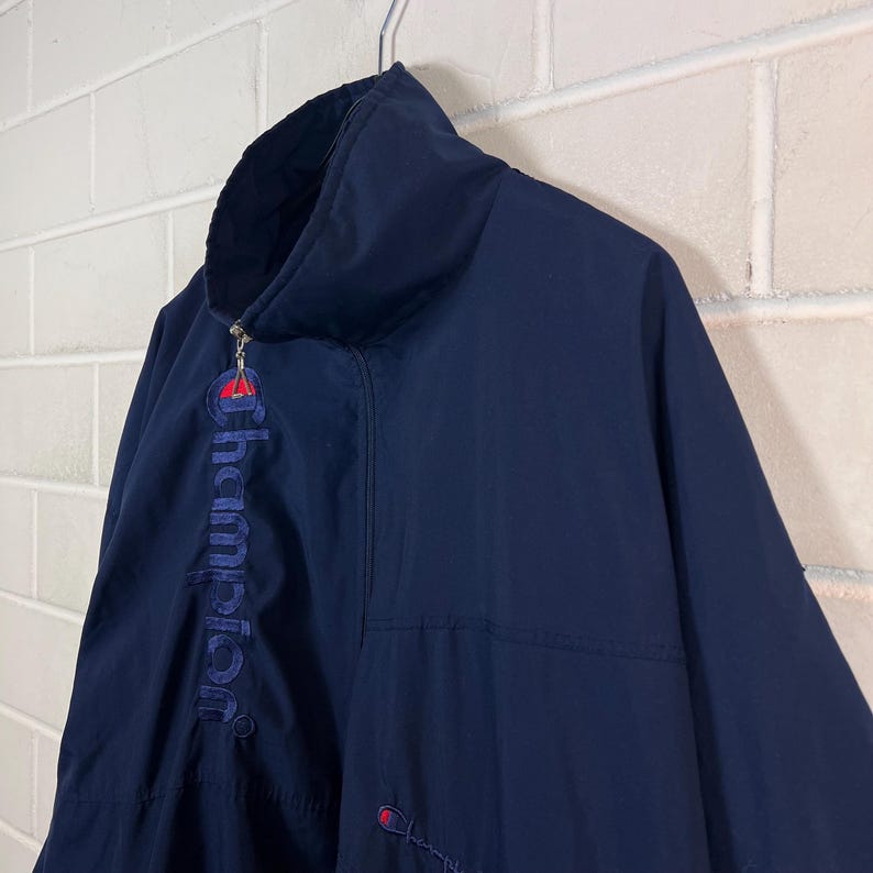 May include: Navy blue Champion jacket with a high collar and a full zipper. The Champion logo is embroidered in red and blue on the chest and the word "Champion" is embroidered vertically on the left side. The jacket is hanging on a metal hanger.