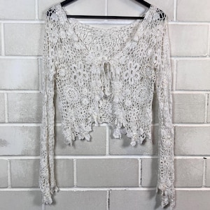 Könnte beinhalten: Cremefarbenes Häkeltop mit langen Ärmeln. Das Oberteil hat einen V-Ausschnitt und einen Bindverschluss an der Brust. Das Häkeldesign umfasst florale und geometrische Muster, die einen zarten und strukturierten Look erzeugen. Das Kleidungsstück hängt an einem schwarzen Kleiderbügel.