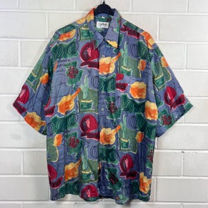 Könnte beinhalten: Ein kurzärmeliges Hemd mit einem farbenfrohen, abstrakten Muster. Das Hemd hat einen hellblauen Hintergrund mit einem sich wiederholenden Design aus Früchten, Flaschen und anderen Formen in Rot, Orange, Grün und Gelb. Es hat einen Kragen und hängt an einem schwarzen Kleiderbügel.