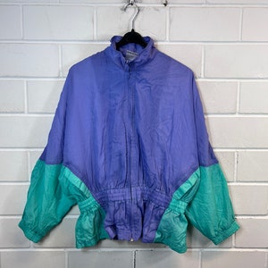 May include: A vintage windbreaker jacket in shades of purple and teal green hangs against a white brick wall. The jacket features a high collar, a full-length zip, and elastic cuffs and waist. The colour-blocked design adds a retro aesthetic.