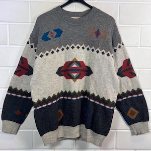 Könnte beinhalten: Ein grauer, cremefarbener und marineblauer Pullover mit geometrischen Mustern. Der Pullover hat einen Rundhalsausschnitt und lange Ärmel. Das Design umfasst Diamanten, Dreiecke und Rechtecke in Rot, Blau und Braun. Der Pullover hängt an einem schwarzen Kleiderbügel.