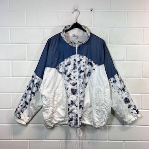 Pode incluir: Uma jaqueta corta-vento branca e azul com um padrão tie-dye. A jaqueta tem um fecho de correr e dois bolsos na frente.