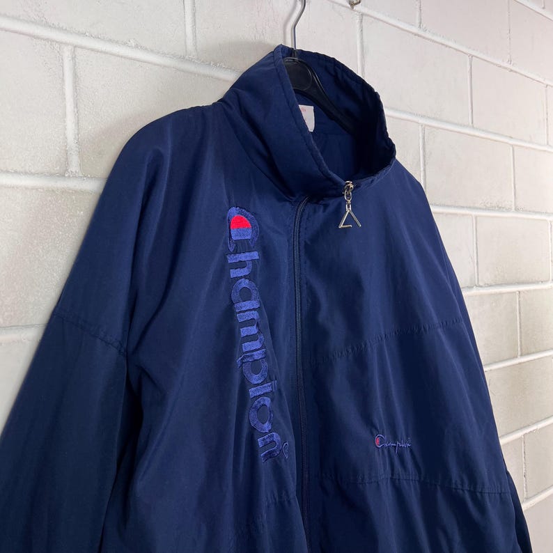 May include: Navy blue Champion zip-up jacket. The jacket features the Champion logo in red and blue on the chest and the word "Champion" vertically embroidered on the left side. The jacket has a high collar and a zipper closure.