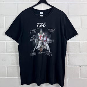 Könnte beinhalten: Schwarzes T-Shirt mit einer Grafik eines Ritters in Rüstung, dem Text "Armor of God" und beschrifteten Komponenten. Der Ritter hat ein Schwert und einen Schild mit einem roten Kreuz auf dem Schild.