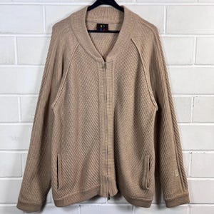 May include: A beige zip-up cardigan with a ribbed texture. The cardigan features a V-neck, two front pockets, and a small logo on the left sleeve. The zip runs down the centre. The cardigan is hanging on a black hanger.