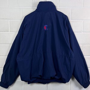 May include: Navy blue Champion jacket with a red and blue logo on the back. The jacket has a high collar, long sleeves, and a drawstring waist. The jacket is made of a lightweight material.