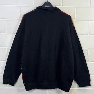 K&ouml;nnte beinhalten: Schwarzer Strickpullover mit Kragen. Die Schultern haben ein Muster in Rot, Braun und Gelb. Der Pullover h&auml;ngt vor einer wei&szlig;en Backsteinmauer.