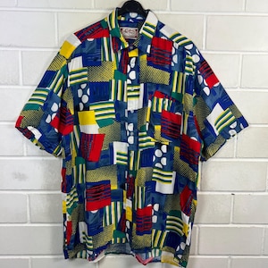 Può includere: Camicia button-down a maniche corte con un vivace design patchwork. La camicia presenta un mix di forme e motivi geometrici blu, rossi, gialli, verdi e bianchi. La camicia è appesa a una gruccia nera contro un muro di mattoni bianchi.