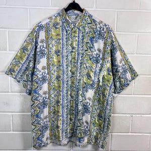 May include: A short-sleeved button-down shirt with a white background and a colorful floral pattern in shades of blue, green, and yellow. The shirt is hanging on a black hanger.