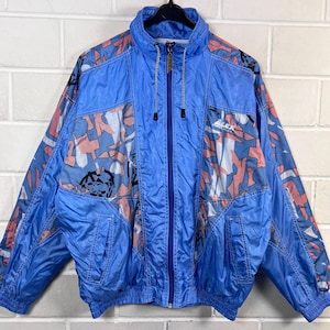 May include: A vintage blue windbreaker jacket with a full zipper closure. The jacket features a geometric pattern in pink, white, and black. It has a high collar and two front pockets. The jacket is made of a shiny, lightweight material.