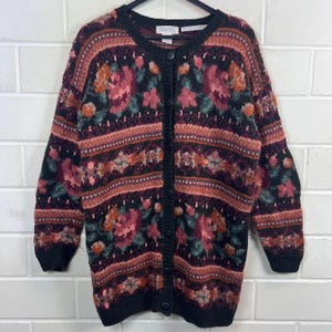 Könnte beinhalten: Vintage schwarze Strickjacke mit floralem Muster. Der Cardigan mit Knöpfen hat rosa, orange und grüne Blumen mit horizontalen Bändern aus geometrischen Designs. Die Ärmel und der Kragen sind schwarz. Das Etikett lautet "Word".