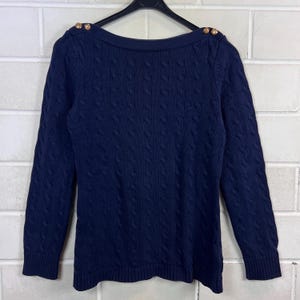 Könnte beinhalten: Ein marineblauer Pullover mit Zopfmuster. Der langärmlige Pullover hat einen U-Boot-Ausschnitt und goldfarbene Knöpfe auf den Schultern. Der Pullover hängt an einem schwarzen Kleiderbügel vor einer weißen Backsteinmauer.