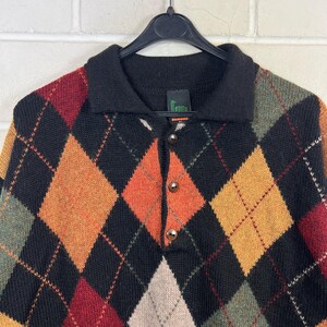 K&ouml;nnte beinhalten: Ein schwarzer Argyle-Pullover mit Kragen und drei Kn&ouml;pfen. Der Pullover hat ein Rautenmuster in Orange, Gelb, Gr&uuml;n und Rot. Der Pullover h&auml;ngt an einem schwarzen Kleiderb&uuml;gel.