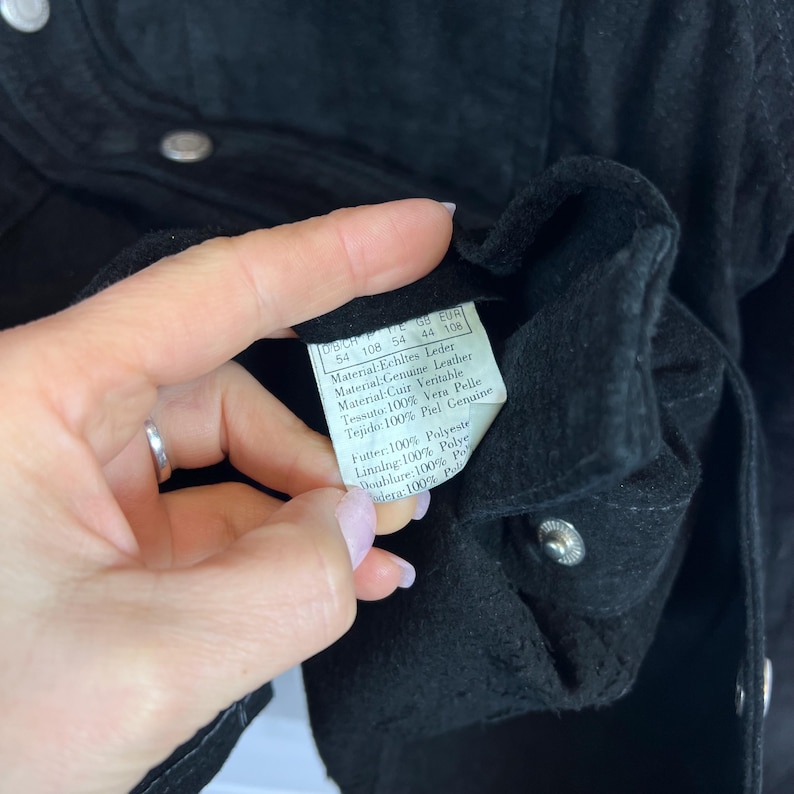 Peut inclure: Gros plan sur une veste en cuir noir avec une &eacute;tiquette. L'&eacute;tiquette indique le cuir v&eacute;ritable et comprend des d&eacute;tails en plusieurs langues. La veste a des fermetures &agrave; pression argent&eacute;es.