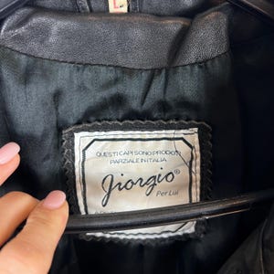 May include: Close-up of a black leather jacket's interior, showcasing a label with the brand name "Giorgio" and Italian text. The label is rectangular with a decorative border.