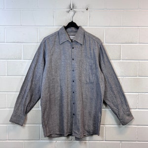 May include: A grey long-sleeved button-down shirt with a pocket on the left side. The shirt is hanging on a black hanger against a white brick wall.
