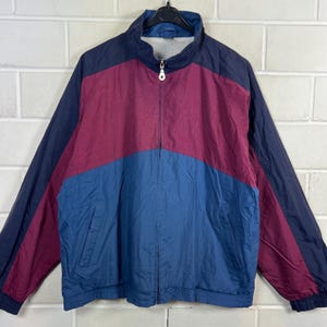 May include: A colour-blocked jacket in navy, burgundy, and blue. The jacket has a full zip, a stand-up collar, and elastic cuffs. The jacket is made of a lightweight material, possibly nylon. The jacket is hanging on a hanger against a white brick wall.
