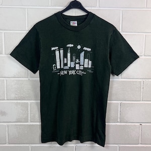 Könnte beinhalten: Ein schwarzes Kurzarm-T-Shirt mit einem weißen Grafikdruck der New York City Skyline, einschließlich der Zwillingstürme, und dem Text "NEW YORK CITY" unter den Gebäuden. Das T-Shirt hat einen Rundhalsausschnitt und eine normale Passform.