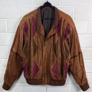 Könnte beinhalten: Eine braune Wildleder-Bomberjacke mit geometrischem Rautenmuster in Dunkelbraun und Burgunderrot. Die Jacke hat einen einfarbigen braunen Kragen, gerippte Bündchen und Saum, eine Knopfleiste vorne sowie ein dunkles Innenfutter mit wiederkehrendem Muster.
