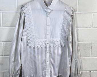 May include: White button-down blouse with vertical stripes and lace detailing. The long-sleeved shirt features a high collar and decorative lace panels on the front. The blouse is hanging on a black hanger against a white brick wall.