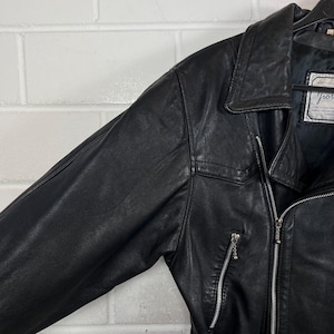May include: Black leather jacket with a notched collar, zipper closure, and a small pocket on the chest. The jacket has a classic design and appears to be in good condition. The jacket has a label with text.