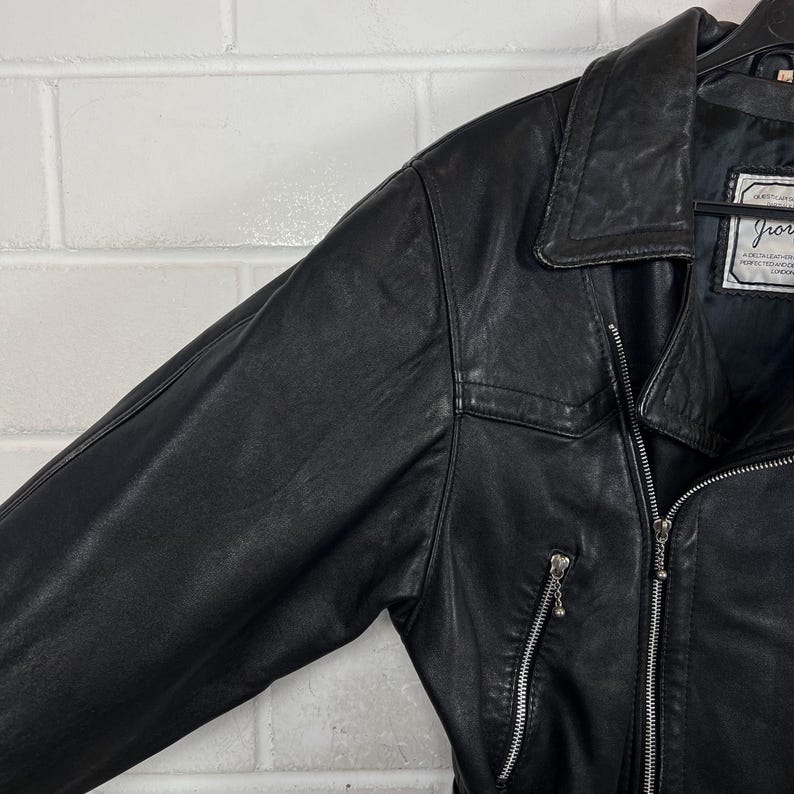 May include: Black leather jacket with a notched collar, zipper closure, and a small pocket on the chest. The jacket has a classic design and appears to be in good condition. The jacket has a label with text.