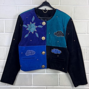Può includere: Una giacca in camoscio a blocchi di colore con maniche nere e un pannello frontale diviso in sezioni blu, viola e verde acqua. La giacca è decorata con motivi ricamati di sole e nuvole, bottoni dorati a forma di stella e piccole borchie dorate.