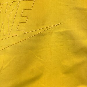 Puede incluir: Primer plano de una tela amarilla con un swoosh Nike bordado en blanco y la palabra "NIKE" en una fuente en negrita y de bloque.