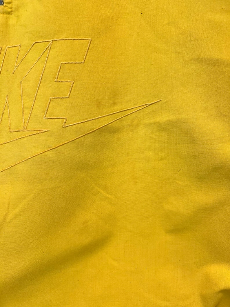 Puede incluir: Primer plano de una tela amarilla con un swoosh Nike bordado en blanco y la palabra "NIKE" en una fuente en negrita y de bloque.