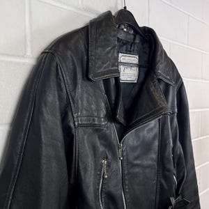 May include: Black leather jacket with a notched collar, asymmetrical zipper, and a small pocket on the chest. The jacket is hanging on a black hanger against a white brick wall. The jacket has a label with text.