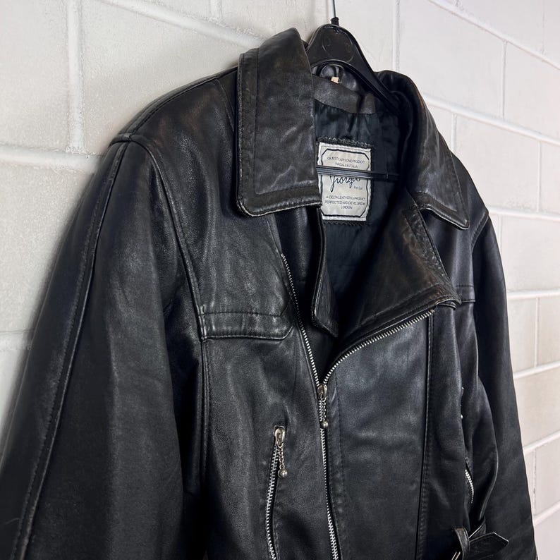 May include: Black leather jacket with a notched collar, asymmetrical zipper, and a small pocket on the chest. The jacket is hanging on a black hanger against a white brick wall. The jacket has a label with text.