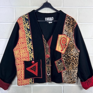 Könnte beinhalten: Eine schwarze Langarm-Strickjacke mit V-Ausschnitt und Knopfleiste. Das Kleidungsstück zeigt eine Mischung aus verschiedenen Mustern, darunter ein rot-goldenes Spiralmuster, ein Leopardenmuster-Abschnitt und ein Aufnäher mit roter Spirale.