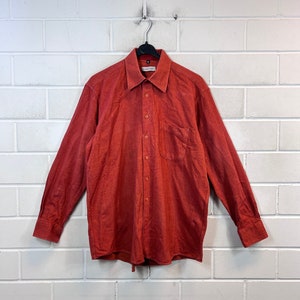 May include: A long-sleeved, button-down shirt in a solid rust orange colour. The shirt has a single chest pocket and a button-down collar.