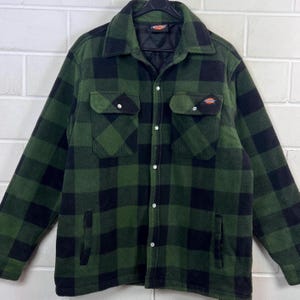 Könnte beinhalten: Grün-schwarze karierte Jacke mit zwei Brusttaschen und zwei Seitentaschen. Die Jacke hat eine Knopfleiste und ein schwarzes Steppfutter. Das Dickies-Logo ist auf einer der Brusttaschen sichtbar.