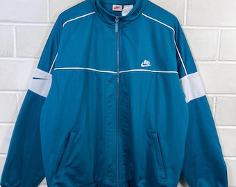 May include: Teal Nike track jacket with white accents. Features a full zip closure, a stand-up collar, and the Nike logo on the chest. White stripes run across the chest and down the sleeves. A classic athletic jacket.