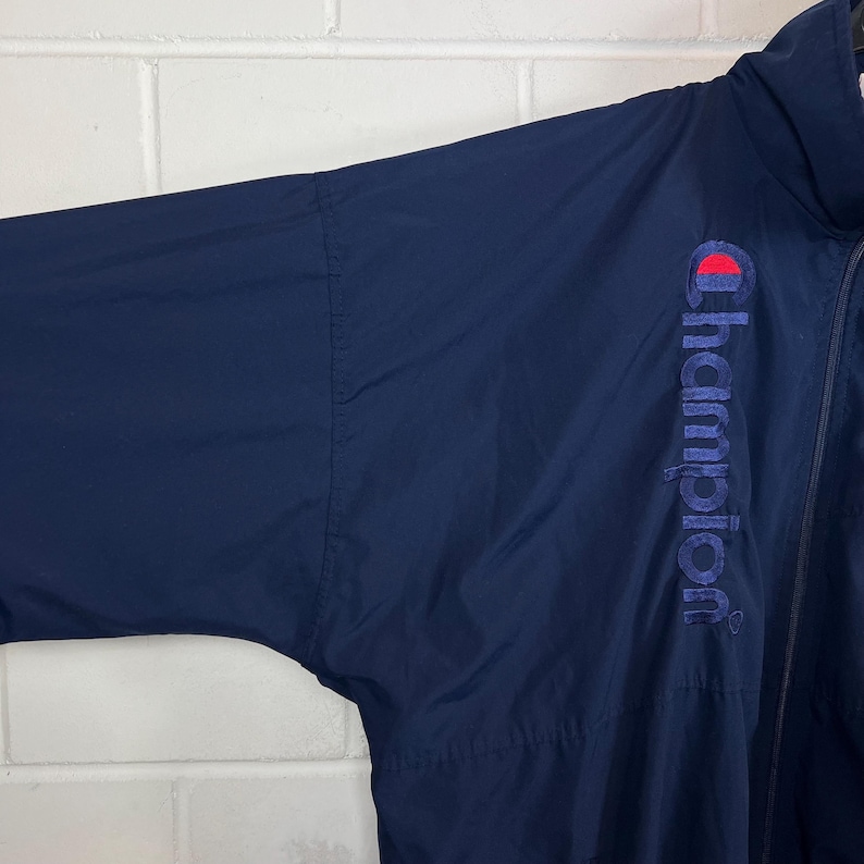May include: Navy blue Champion jacket with a full zipper. The Champion logo is embroidered in red and white on the upper right sleeve. The jacket appears to be made of a lightweight, water-resistant material.