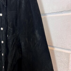 Peut inclure: Veste en daim noire avec des boutons blancs et une poche poitrine. La veste a un col classique et une coupe ajust&eacute;e. Le daim a une texture douce et velout&eacute;e.