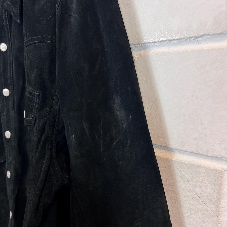 Peut inclure: Veste en daim noire avec des boutons blancs et une poche poitrine. La veste a un col classique et une coupe ajust&eacute;e. Le daim a une texture douce et velout&eacute;e.