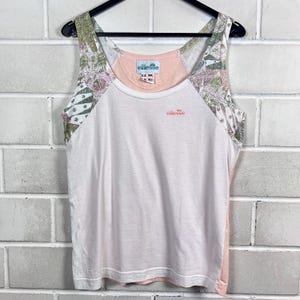 Könnte beinhalten: Ein weißes und rosa Tanktop mit einem Blumenmuster auf den Schultern und einem kleinen Logo auf der Brust. Das Tanktop hängt an einem schwarzen Kleiderbügel vor einer weißen Backsteinmauer.