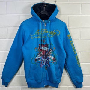 May include: A turquoise zip-up hoodie with a black interior hood. The front features a skull and rose graphic with the words "Love Kills Slowly." The Ed Hardy logo is in gold script across the chest, with "By Christian Audigier" printed on the sleeve.